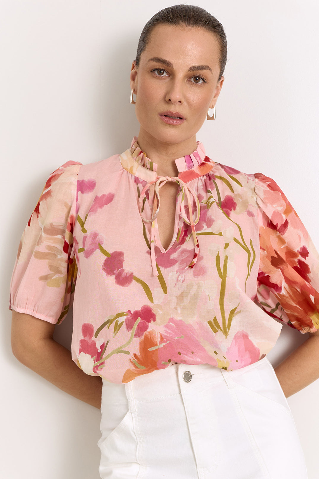 A woman wearing a light pink floral blouse with puff sleeves, a tie-neck detail, and a watercolor-style print in pink, red, and green, paired with white pants against a plain light background.