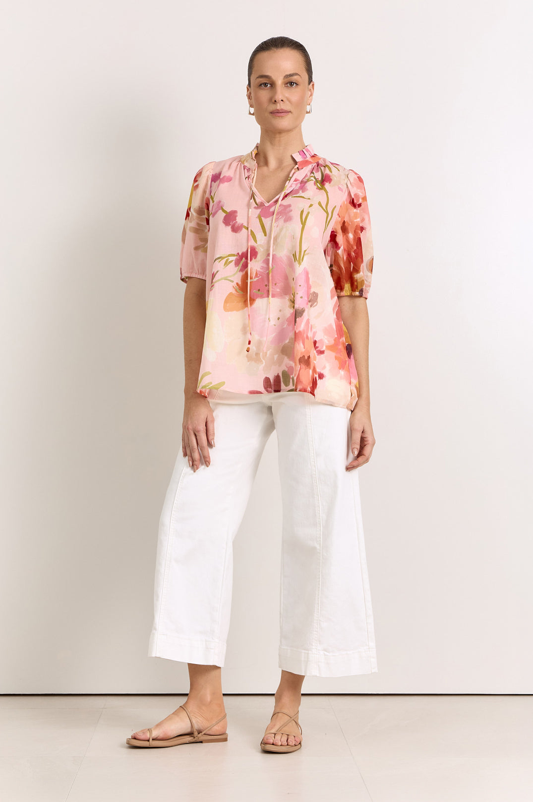 A woman wearing a short-sleeve blouse with a pink, red, and orange floral print, paired with white cropped pants and flat sandals, standing against a plain off-white studio background.