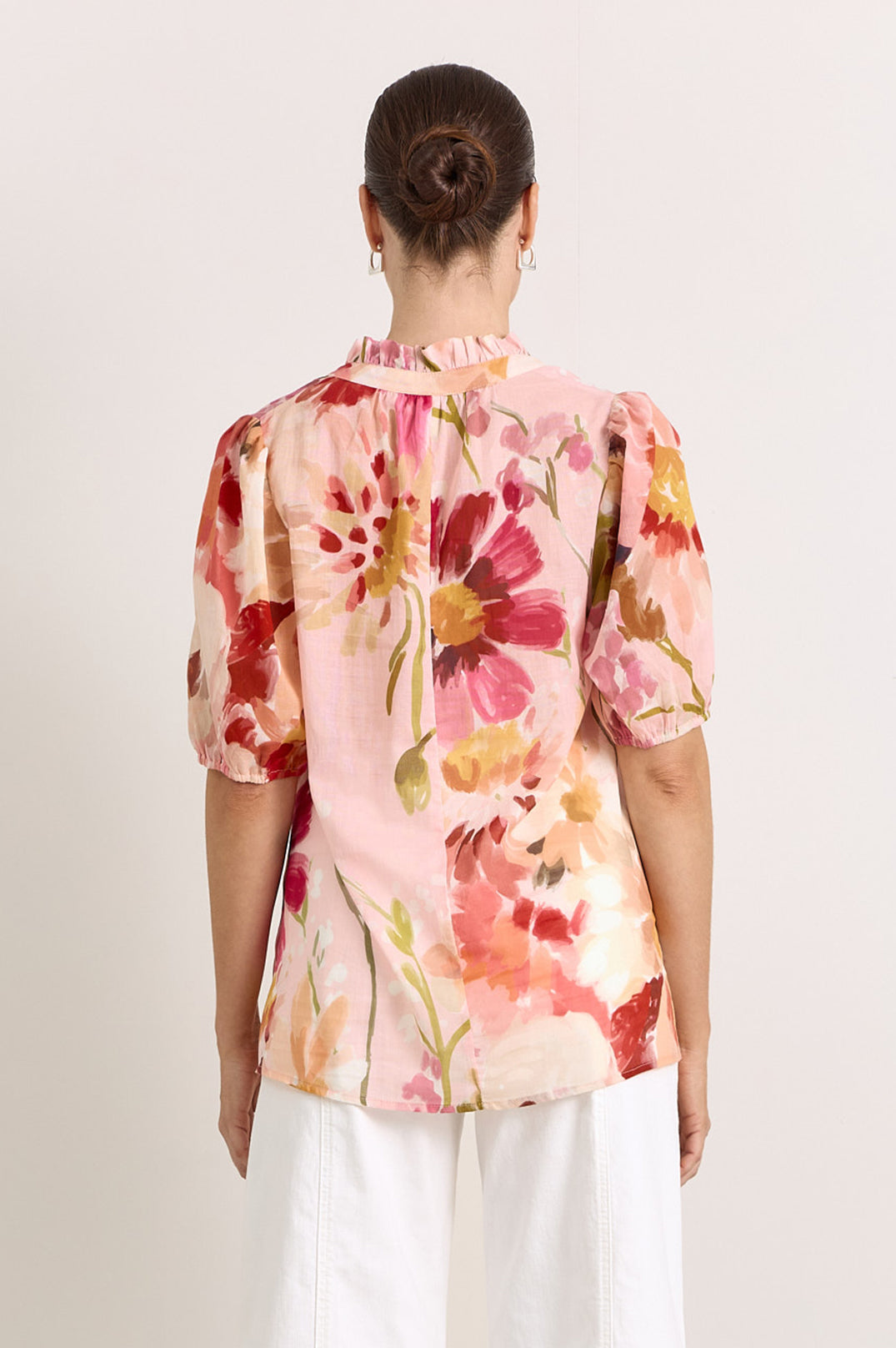 Image of A woman viewed from the back wearing a light pink short-sleeve blouse with a large watercolor-style floral print in red, orange, and yellow, paired with white pants, against a plain light background.