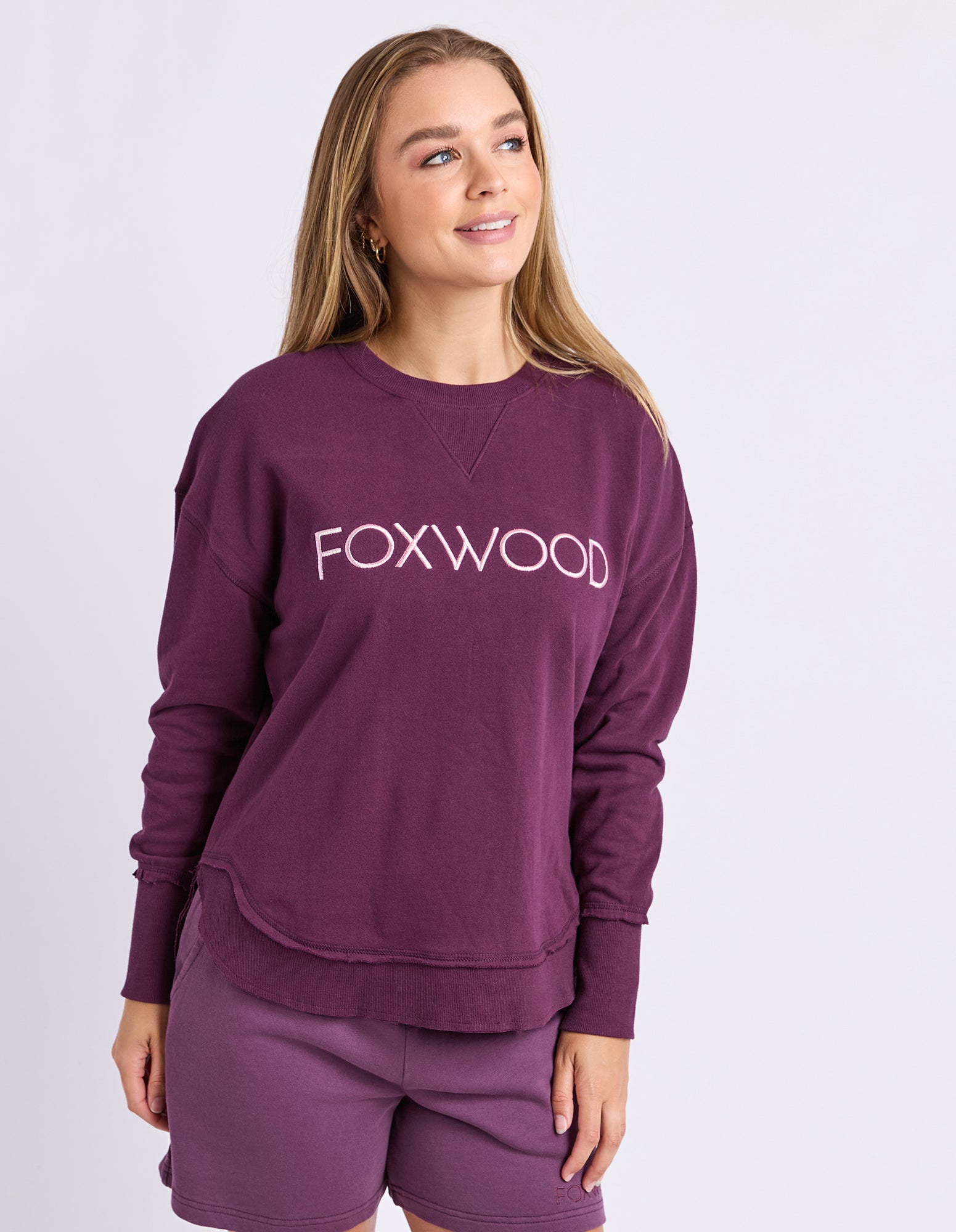 Image of A woman wearing a plum Foxwood jumper and matching shorts, standing against a light grey studio background.
