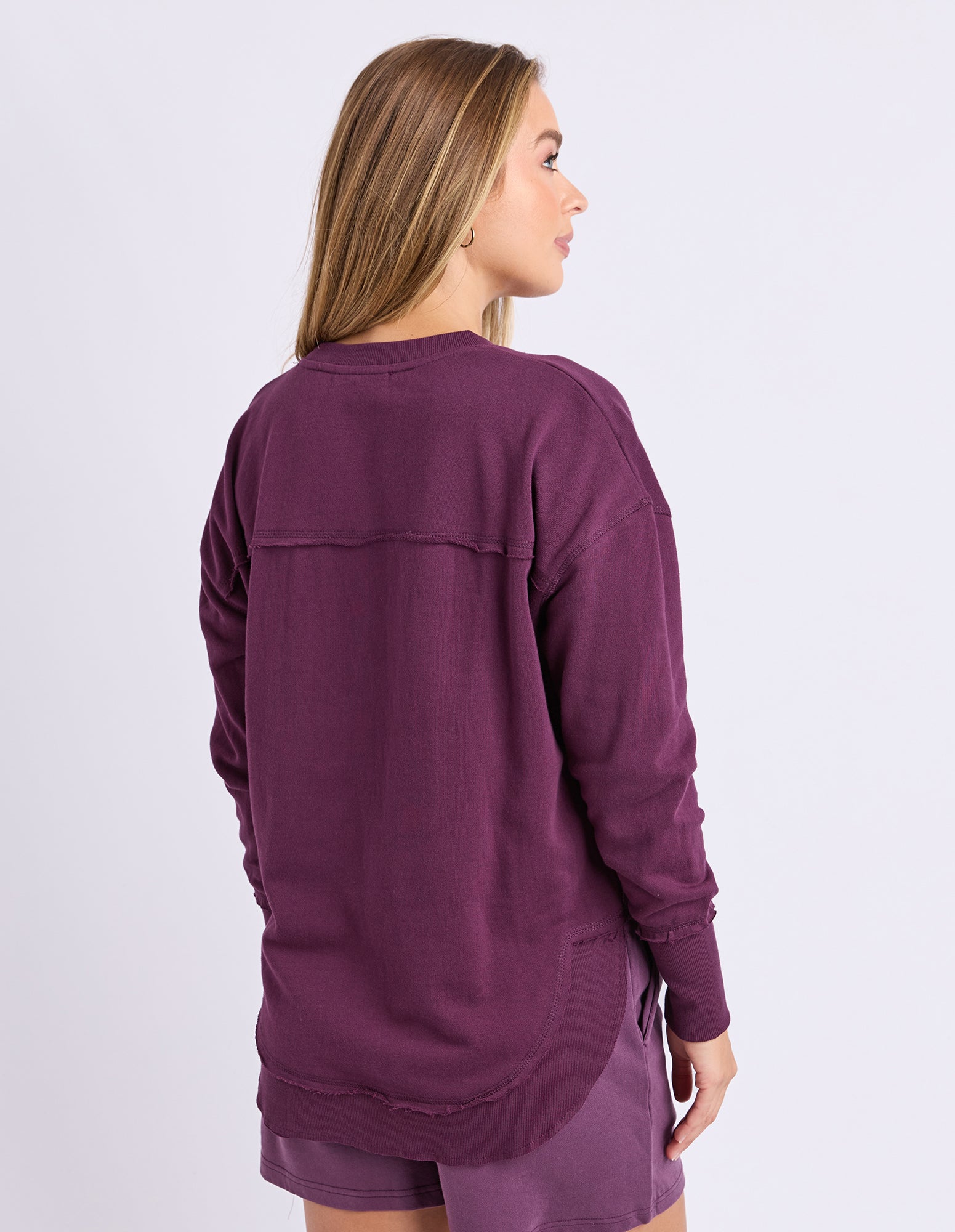 A woman shown from the back wearing a long-sleeve crewneck jumper in a plum purple coloru, paired with matching shorts, against a light grey studio background.