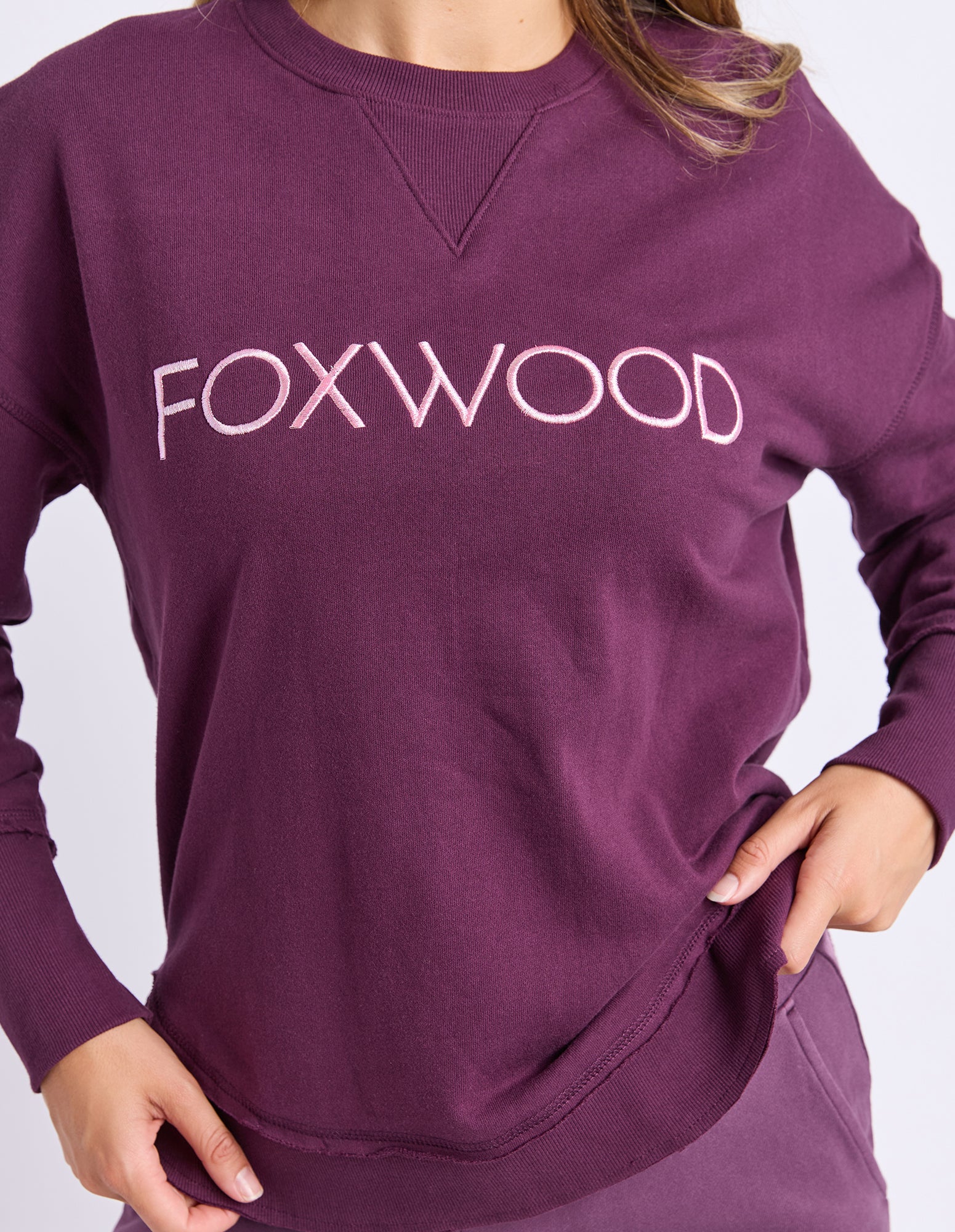 Image of A close-up of a woman wearing a long-sleeve crewneck jumper in deep plum purple with the brand name 'FOXWOOD' printed across the chest in a light pink font.